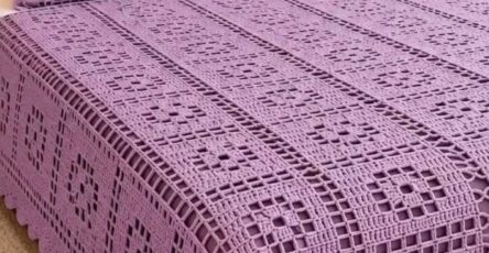 A lavender crochet bedspread and matching pillows with openwork square motifs and a scalloped border on a neatly made bed.