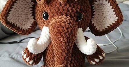 Cute crocheted plush mammoth amigurumi with big floppy ears, curly hair, and white tusks, made from soft brown and white yarn.