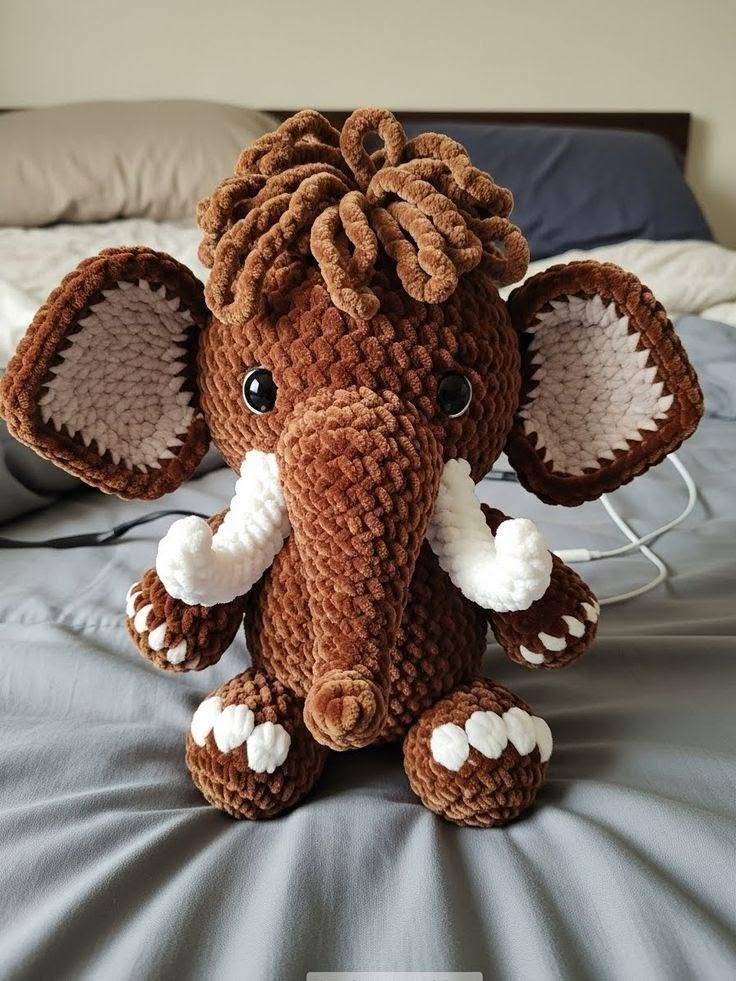 Cute crocheted plush mammoth amigurumi with big floppy ears, curly hair, and white tusks, made from soft brown and white yarn.