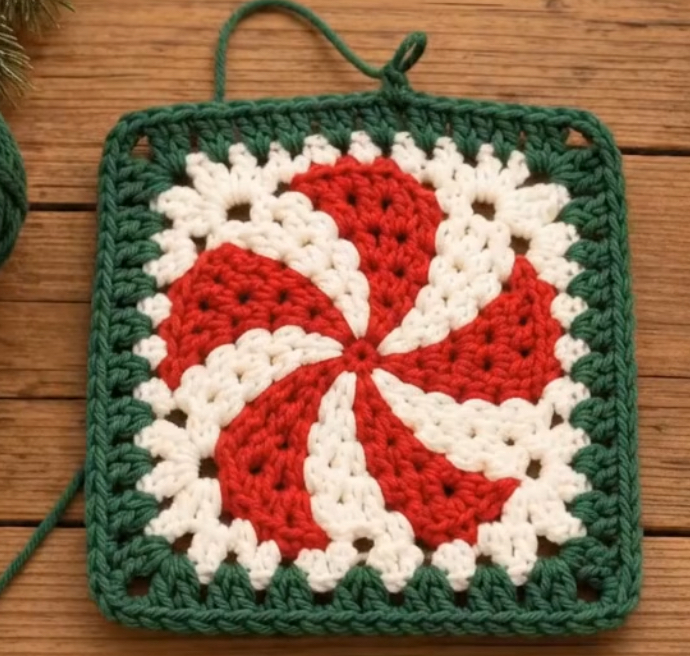 Crocheted square featuring a red and white swirl motif resembling a peppermint candy with a green granny square border.