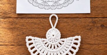 A delicate white crochet angel ornament with detailed lace patterning, displayed on a wooden surface, next to a drawn crochet diagram.