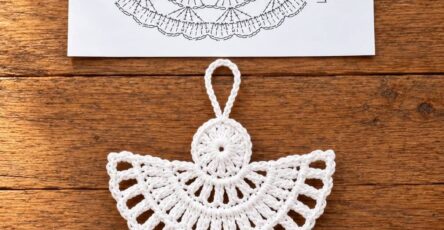 A delicate white crochet angel ornament with detailed lace patterning, displayed on a wooden surface, next to a drawn crochet diagram.