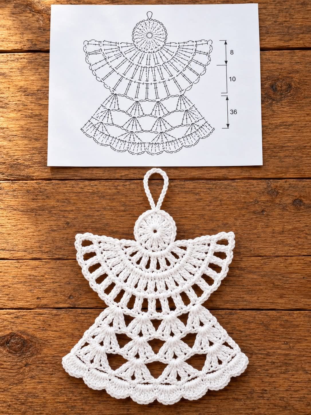 A delicate white crochet angel ornament with detailed lace patterning, displayed on a wooden surface, next to a drawn crochet diagram.