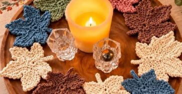 Several hand-crocheted maple leaf motifs in cream, blue, green, and brown tones arranged as autumn-inspired coasters or decor on a wooden tray.
