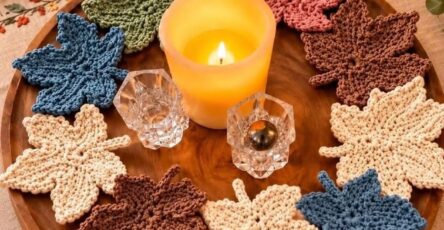 Several hand-crocheted maple leaf motifs in cream, blue, green, and brown tones arranged as autumn-inspired coasters or decor on a wooden tray.