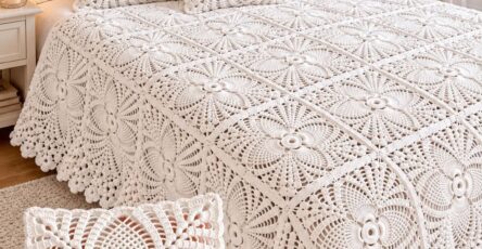 Hand holding a crocheted white lacy floral square with a matching white crochet bedspread and pillows made of similar floral motifs on a bed.