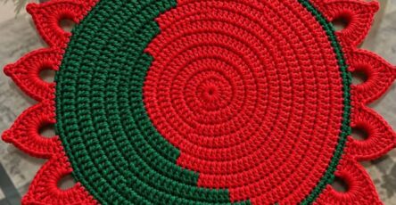 Round crochet placemat with alternating red and green color blocking and petal-shaped edging, displayed on a glass table.