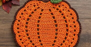 A handmade crochet pumpkin placemat with an orange body, brown stem, and a green leaf on a wooden background.