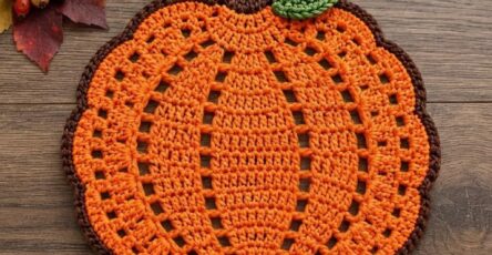 A handmade crochet pumpkin placemat with an orange body, brown stem, and a green leaf on a wooden background.