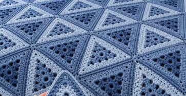 A crocheted blanket made of blue and light blue triangle motifs joined to form a geometric design, displayed on a neatly made bed.