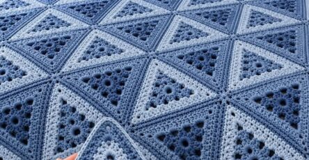 A crocheted blanket made of blue and light blue triangle motifs joined to form a geometric design, displayed on a neatly made bed.