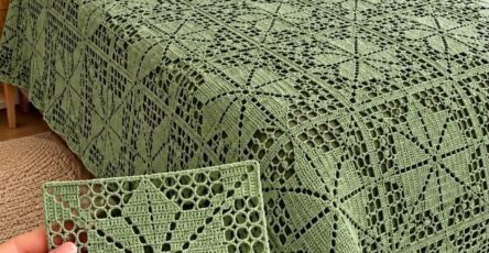 A green crochet bedspread and pillow set made from joined geometric lace squares, with a hand holding a single square motif in the foreground.