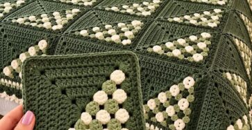 A crocheted blanket and matching pillows with green and cream diagonal bobble granny squares and scalloped border, shown on a bed.