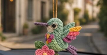 A colorful crocheted hummingbird amigurumi ornament with a flower and leaves, hanging from a car mirror.