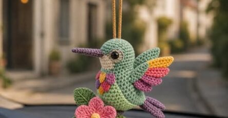 A colorful crocheted hummingbird amigurumi ornament with a flower and leaves, hanging from a car mirror.