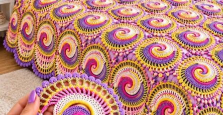 A colorful crochet bedspread made of joined swirling round motifs in purple, yellow, pink, and cream, with decorative pillows and floral accents.