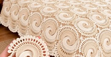 A hand holding a spiral crochet motif in front of a bed covered with a cream-colored crochet spiral motif bedspread.