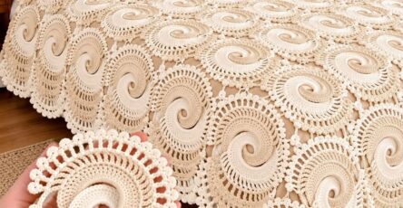 A hand holding a spiral crochet motif in front of a bed covered with a cream-colored crochet spiral motif bedspread.