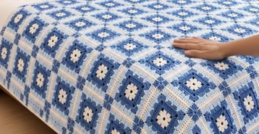 Crocheted blue and white bedspread with flower motif granny squares on a neatly made bed in a bright room.