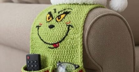 A crochet armrest organizer with a green Grinch face, striped red and white pockets, and a Santa hat on the arm of a couch.