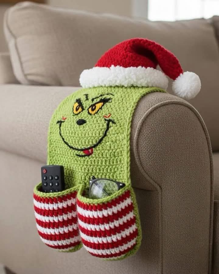 A crochet armrest organizer with a green Grinch face, striped red and white pockets, and a Santa hat on the arm of a couch.