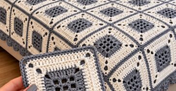 A bed covered with a handmade crochet blanket made from gray and cream granny squares, and a hand holding one finished square in the foreground.