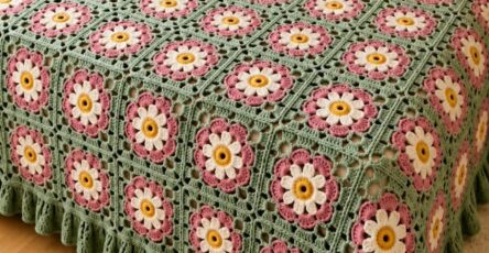 A crochet blanket and matching pillows made from green, pink, white, and yellow floral granny squares on a bed, with a hand holding one square.