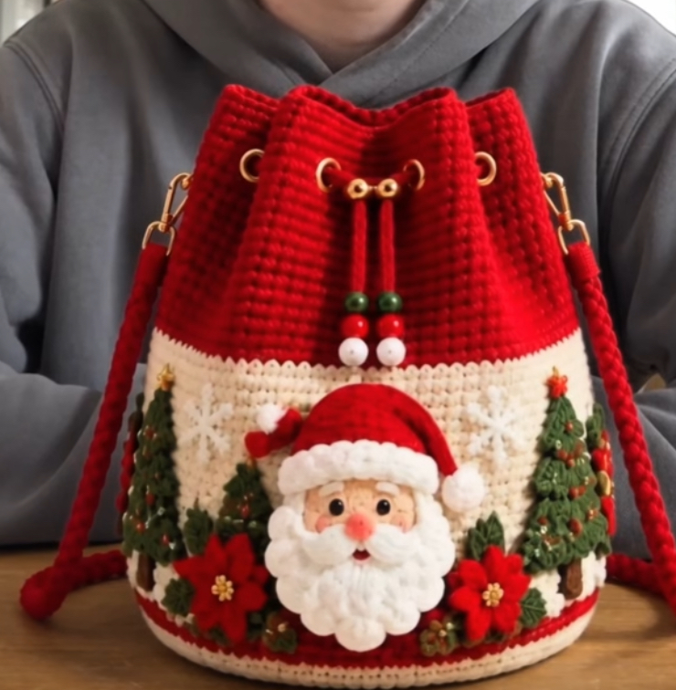 A festive crochet bucket bag with red top, cream body, 3D Santa face, Christmas trees, poinsettias, and drawstring closure.
