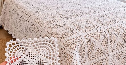 A hand holds a single white lacy crochet square motif in front of a bed covered by a matching intricate crochet bedspread and two crochet pillows.