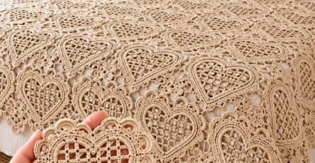 A beige crochet bedspread and pillows with intricate lacy heart motifs arranged in a grid pattern, shown on a neatly made bed.