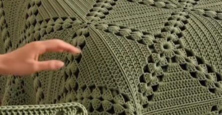 A hand holding a textured green crochet square, part of an olive-colored crocheted bedspread made of joined squares on a bed.