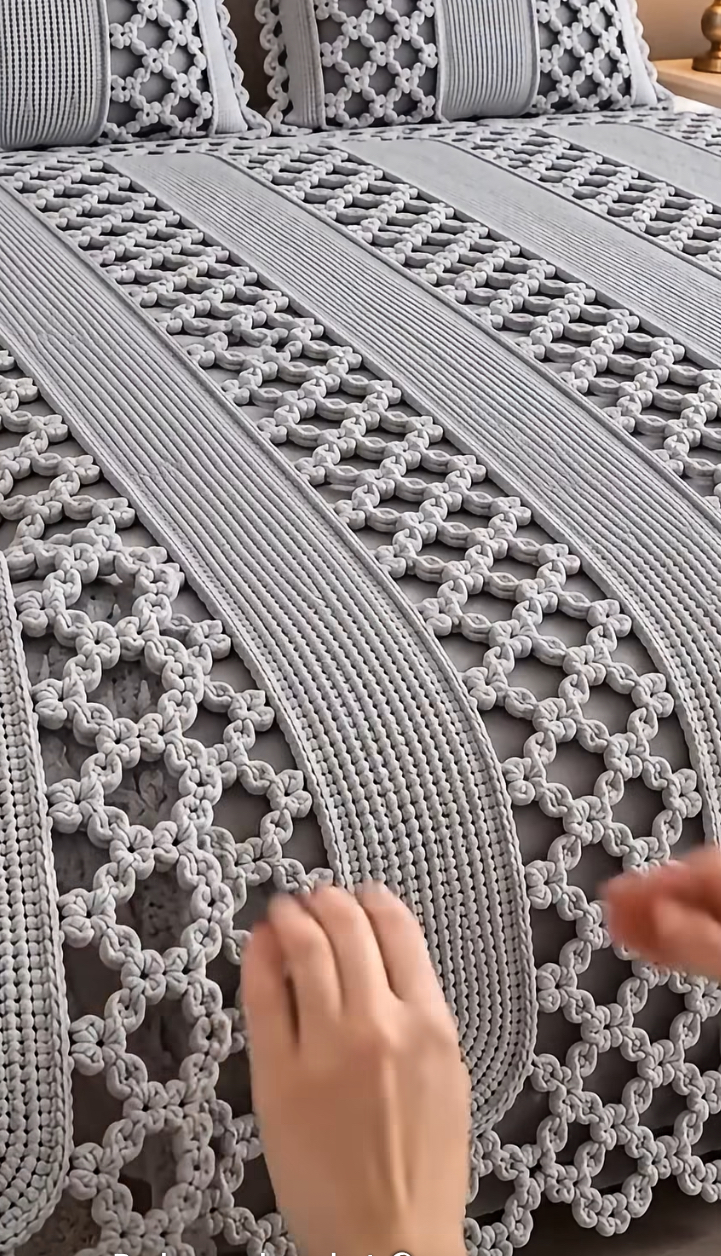A modern crochet bedspread with alternating solid textured stripes and open lattice panels in soft gray yarn, draped smoothly over a bed.