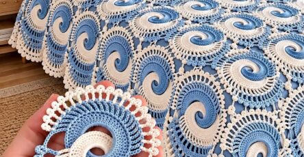 A hand holds a blue and white spiral crochet motif in front of a bed covered with similar swirl motifs forming a decorative crocheted bedspread.