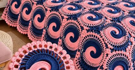 A crochet blanket made from joined pink and navy spiral motifs with decorative openwork edging, spread on a bed.