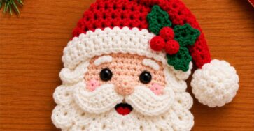 A crocheted Santa Claus face ornament with a red hat, white beard, black eyes, green holly leaves, and red berries, displayed on a wooden surface.