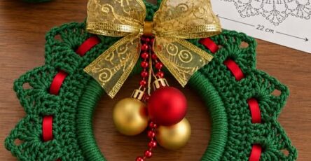 A crocheted green Christmas wreath with a scalloped edge, red ribbon, gold bow, and holiday ornaments on a wooden surface.