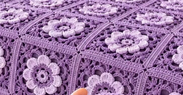 A lavender and white crochet blanket made of joined floral granny squares, with a close-up of a hand holding one square.