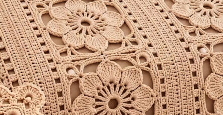 A crocheted bedspread with large floral motifs joined by lacy panels, in a soft beige yarn, draped over a bed with matching pillow covers.