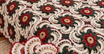 A bed covered with an intricate crochet bedspread featuring red, green, and cream floral motifs, with matching pillows and a hand holding one motif.