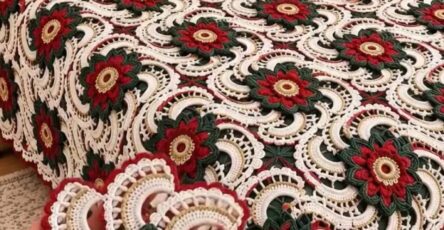 A bed covered with an intricate crochet bedspread featuring red, green, and cream floral motifs, with matching pillows and a hand holding one motif.