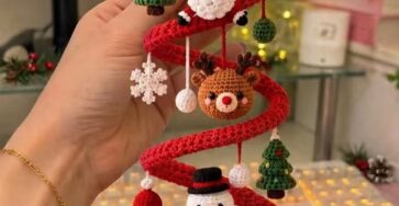 A hand holds a red spiral crochet ornament with attached amigurumi figures, including a star, Santa, reindeer, snowman, gingerbread man, Christmas trees, and snowflakes.