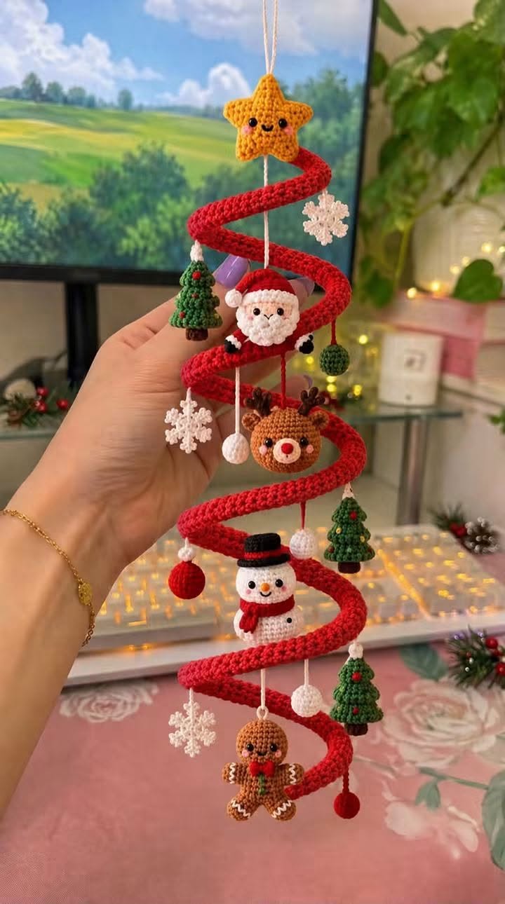 A hand holds a red spiral crochet ornament with attached amigurumi figures, including a star, Santa, reindeer, snowman, gingerbread man, Christmas trees, and snowflakes.