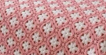 A crochet bedspread with interlocking pink and white daisy granny squares covering a bed, and a hand holding one finished square.