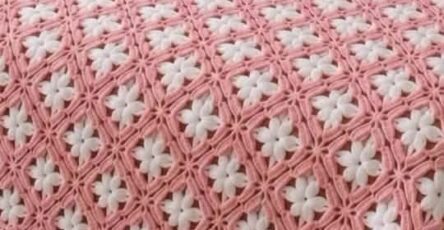 A crochet bedspread with interlocking pink and white daisy granny squares covering a bed, and a hand holding one finished square.