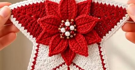 A crochet Christmas star wall hanging in red and white, featuring a poinsettia flower, bow, pine sprigs, and dangling crocheted trees, heart, and bells.