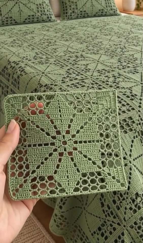 A hand holding a green rectangular crochet motif with a lacy star design, matching a larger bedspread and cushions featuring the same pattern.