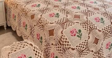 A crochet bedspread made from square motifs with filet rose designs and lacy borders, displayed on a bed in a cozy bedroom.