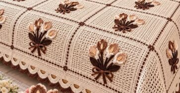 A handmade crochet bedspread with lacy granny squares, brown and beige floral bouquet appliqués, and matching pillows in a rustic bedroom setting.