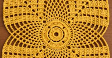 Bright yellow crochet doily with a square shape and radiating petal motifs, displayed with yarn, a crochet hook, and pearls.