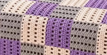 A bed-sized crochet blanket made of purple, gray, and cream block squares in a geometric patchwork design, draped over a bed.